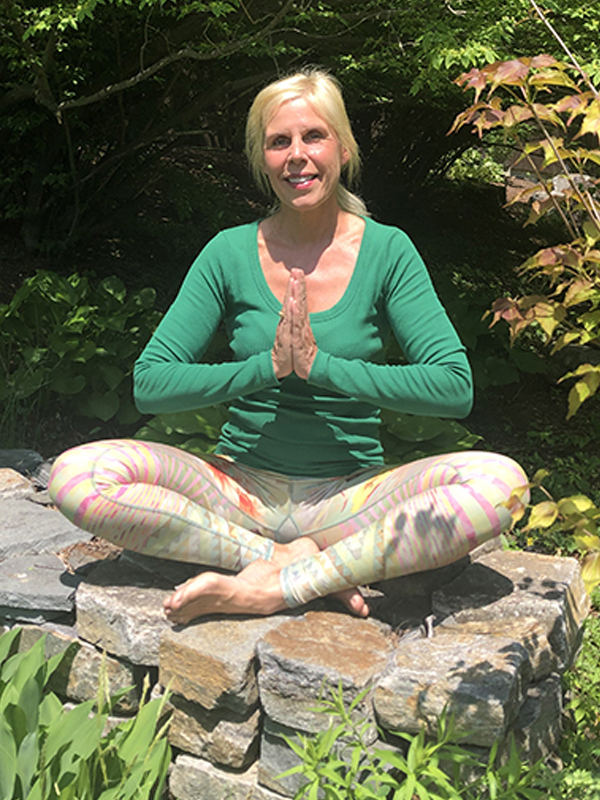 Kim Nashed sitting in lotus position, her hands clasped in a namaste greeting, among the lush foliage of her backyard